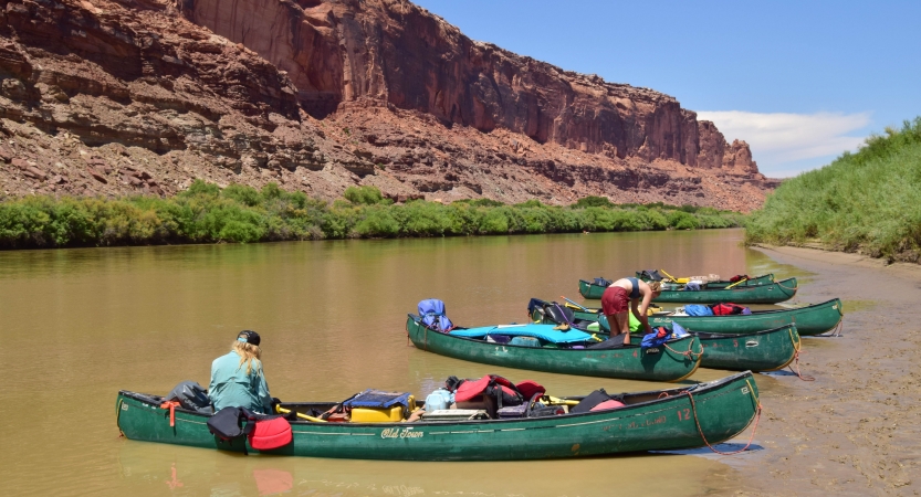 Five canoes rest on the shore of a river. A couple of people are getting things out of the canoes. The other side of the river is lined by a tall red canyon.